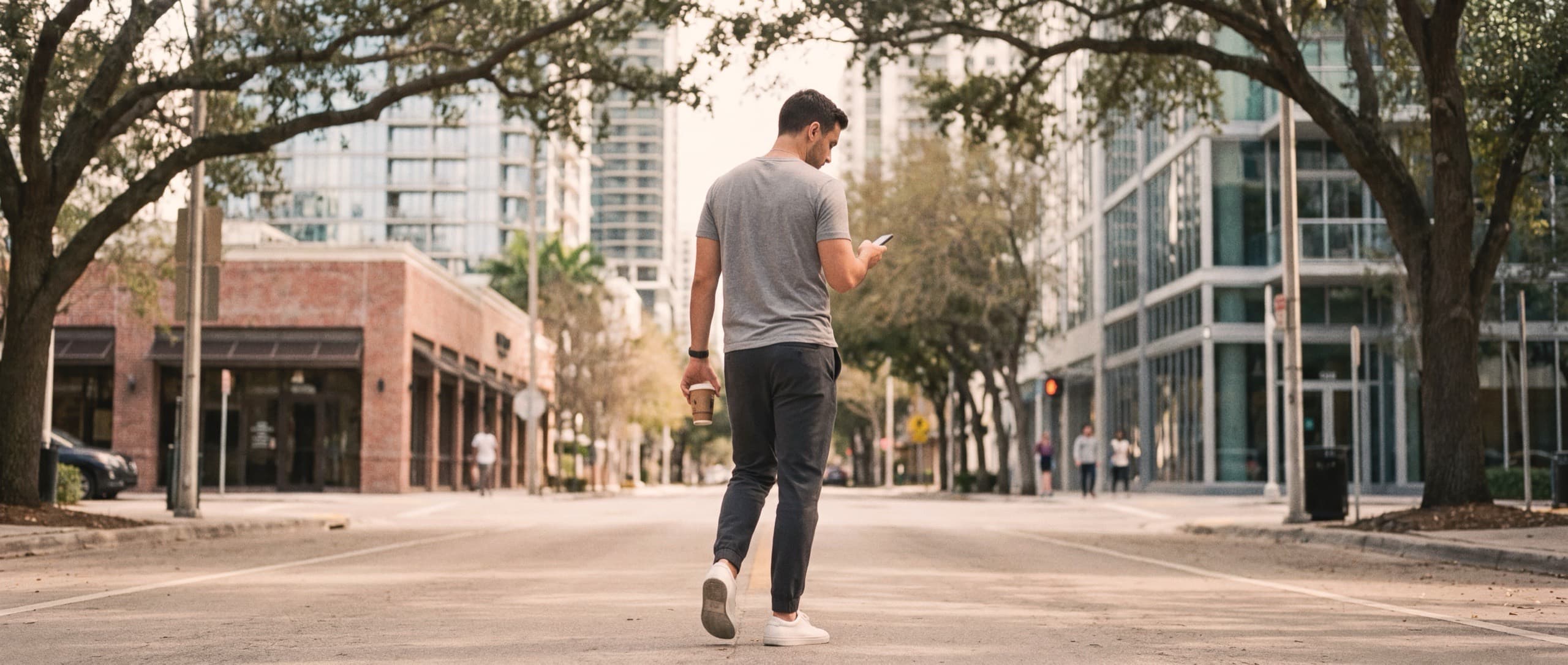 A man seen from behind, face not visible, walking mid-stride along a calm tree-lined downtown Miami street at midday, glancing at a phone held in one hand and carrying a small cafecito cup in the other, two pedestrians at a distance, shot in the desaturated house grade with warm colour and visible film grain