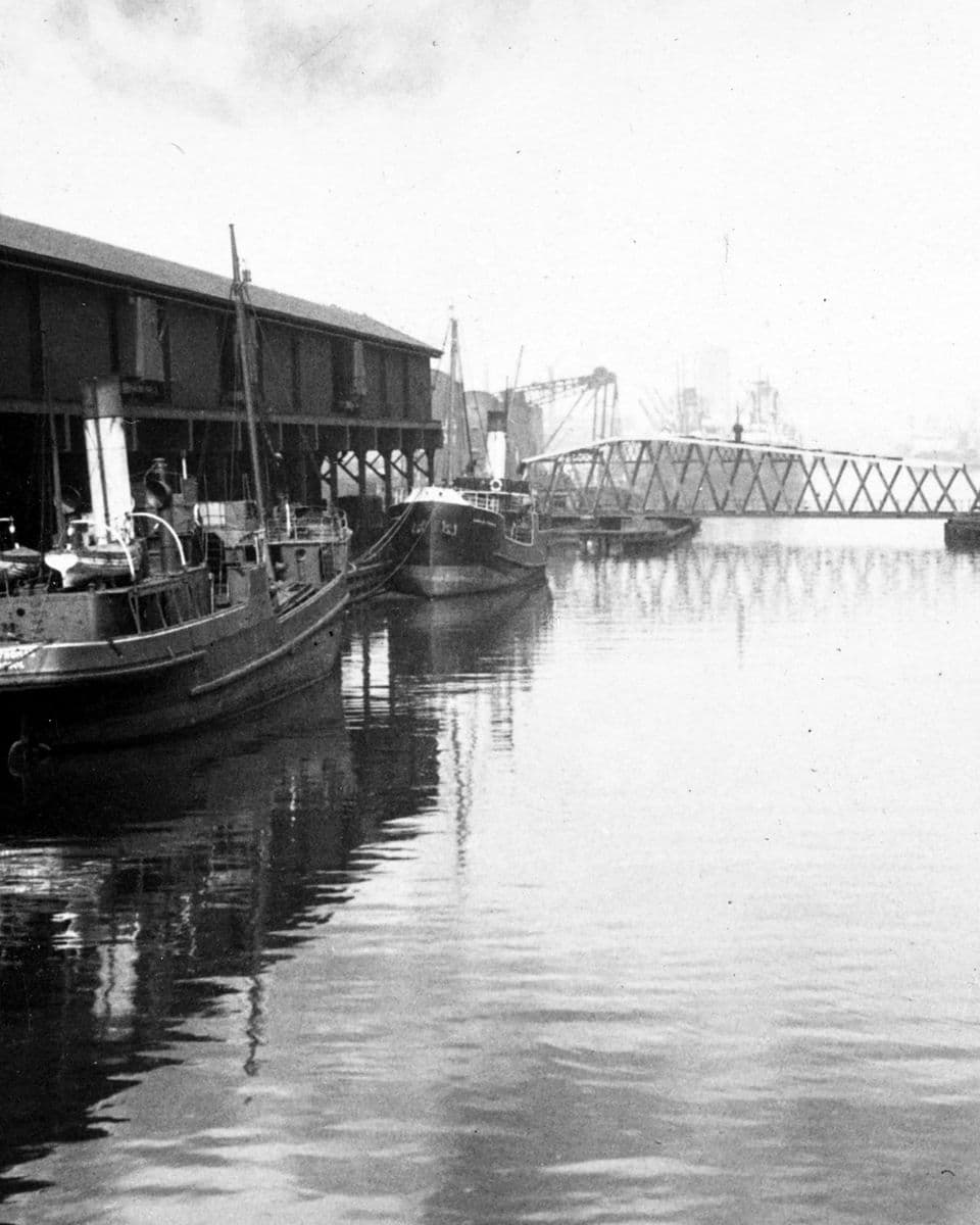 The Manchester Ship Canal in 1928: two steamboats moored at a dockside warehouse, with a long metal bridge beyond.