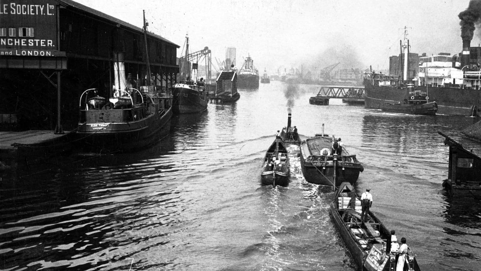Traffic on the Manchester Ship Canal, 1927: a steam tug and barges pass a dockside warehouse, with steamers beyond.